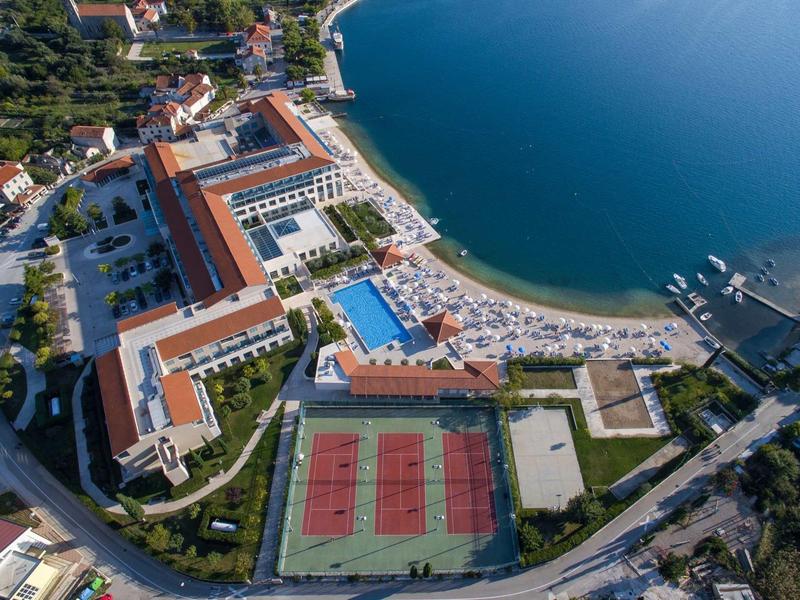 Vista aerea di un resort costiero con campi da tennis, piscine, spiaggia e edifici con tetti rossi.