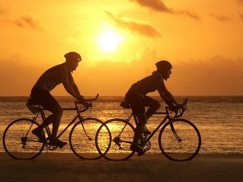 Due persone in bicicletta lungo la spiaggia al tramonto con il sole basso all'orizzonte.