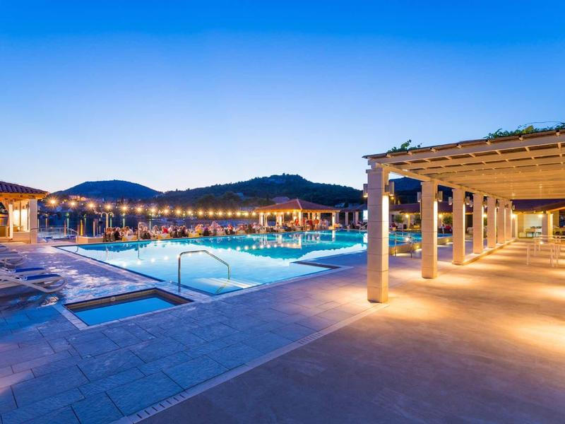 Vista serale di una piscina illuminata con sedie a sdraio e un portico coperto.