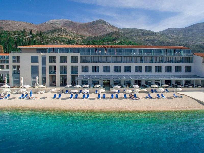 Hotel fronte mare con sdraio e ombrelloni vicino al mare azzurro e montagne.