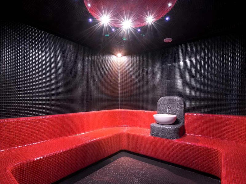 Stanza spa moderna con sedute rosse e sedia grigia a secchiello sotto luci al soffitto luminose.