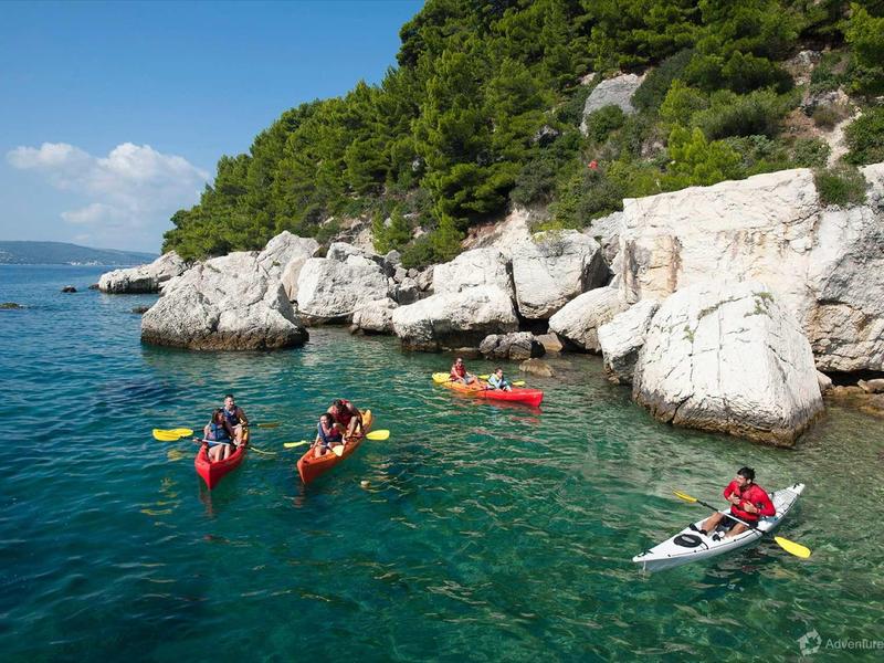 Persone in kayak su acqua blu limpida vicino a costa rocciosa con vegetazione sotto cielo blu.