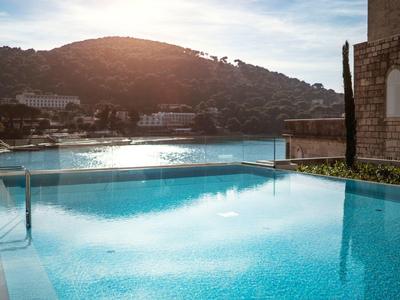 Ein klarer Pool mit Glaszaun vor einem bewaldeten Hügel und Gebäuden im Hintergrund.