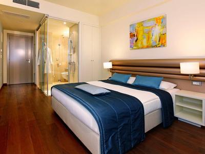 Habitación de hotel moderna con cama doble, suelo de madera y ducha de cristal al fondo.