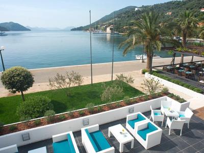 Terrasse mit weißen Möbeln und blauen Kissen, Blick auf Strand, Meer und grüne Hügel im Hintergrund.