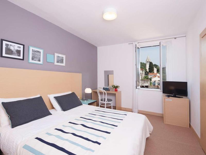 Jasny pokój hotelowy z łóżkiem podwójnym, stolikami nocnymi, biurkiem, telewizorem i oknem z widokiem na miasto.