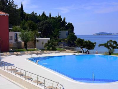 Großer blauer Pool neben roten und weißen Gebäuden mit Meer und bewaldeten Hügeln im Hintergrund.