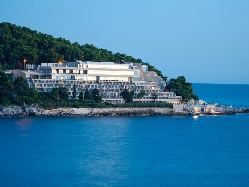 Un gran hotel junto al mar, rodeado de rocas y bosque verde bajo un cielo azul.