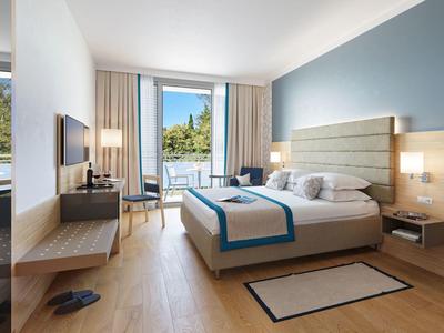 Modernes Hotelzimmer mit Doppelbett, Holzfußboden, großem Fenster und Balkon mit Blick ins Grüne.