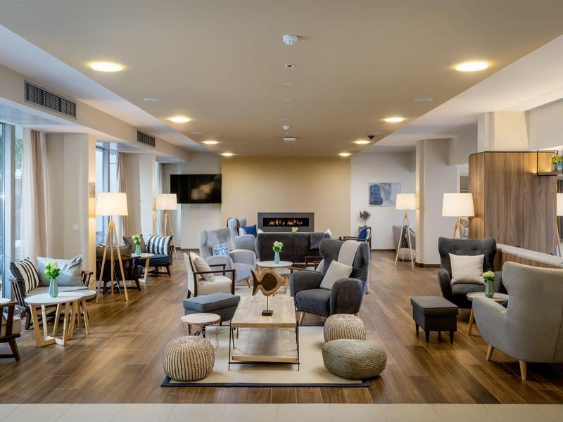 Heller moderner Wohnraum mit Sofas, Sesseln, Holzboden und großen Fenstern, Lampen und Pflanzen.