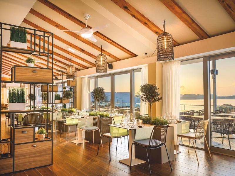 Helles Restaurant mit Holzboden, großen Fenstern, grünen Stühlen und Blick auf Wasser bei Sonnenuntergang.