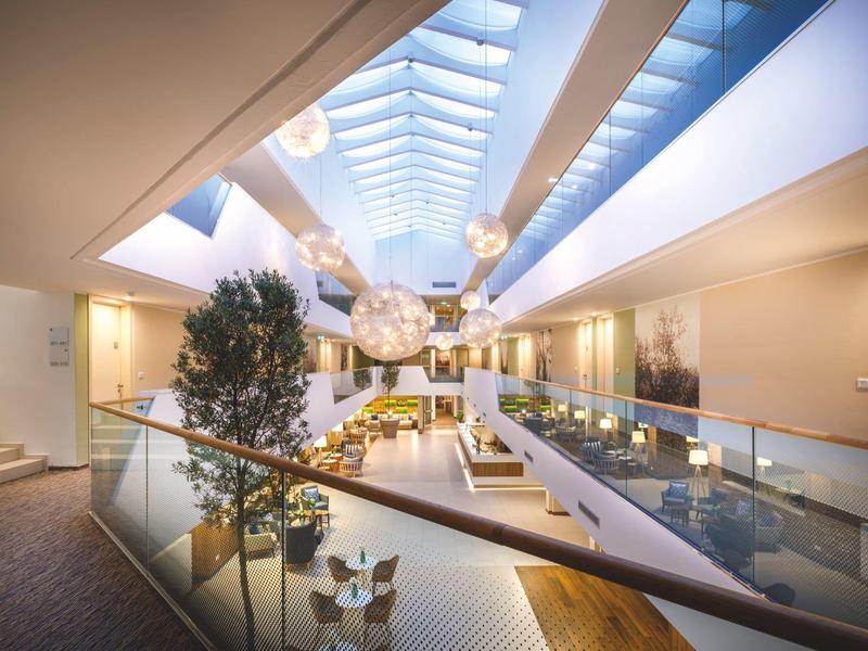 Modernes Atrium mit Glasdach, Pflanzen, Sitzbereichen und hellem, offenem Raumdesign.