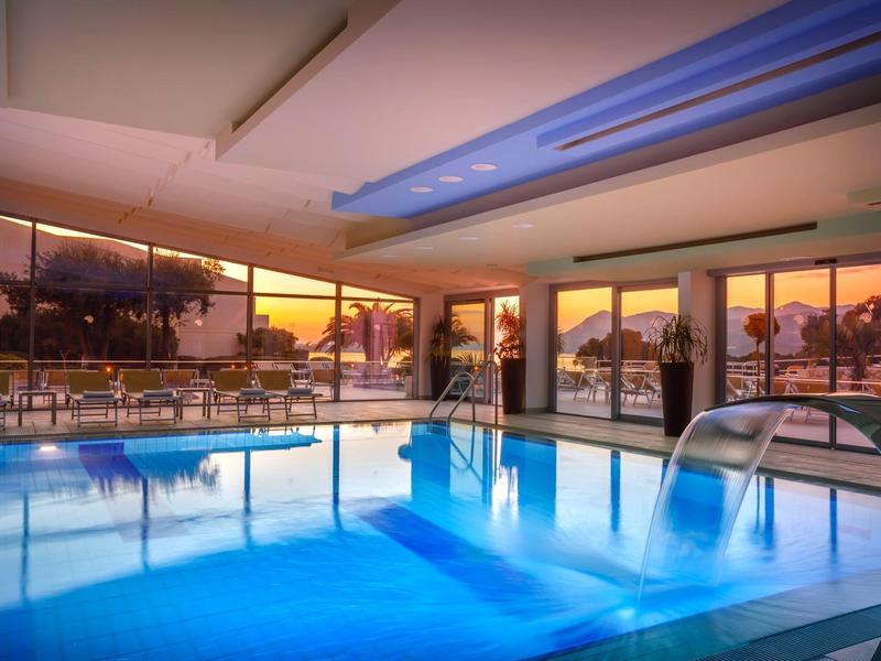 Modernes Hallenbad mit blauer Wasserbeleuchtung und großen Fensterfronten zum Sonnenuntergang.