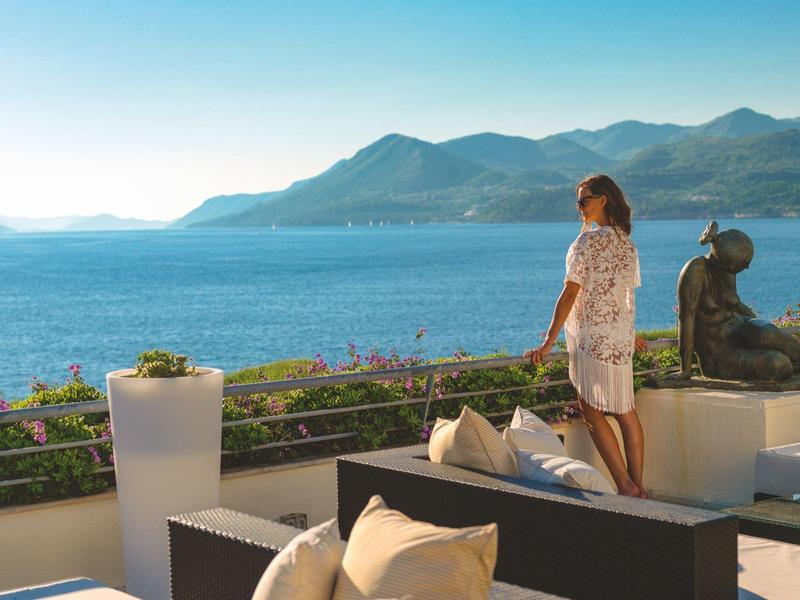 Žena na hotelové terase s výhledem na moře a hory za slunečného dne.
