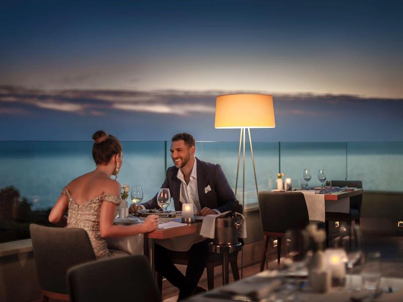 Pár si vychutnává večeři na terase s výhledem na moře za soumraku.