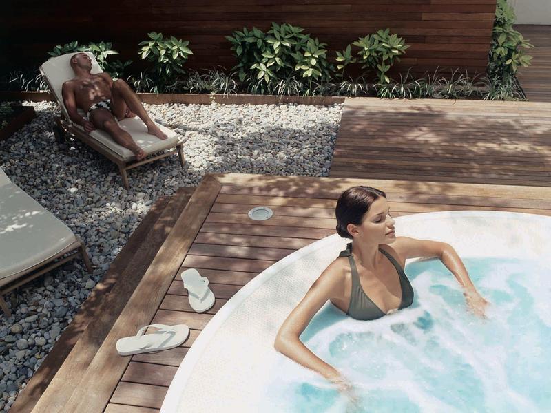 Žena relaxuje ve vířivce na terase, muž leží na lehátku na zahradě.