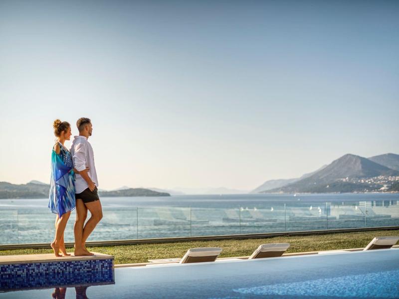 Pár kráčí u bazénu s výhledem na moře a hory v pozadí.