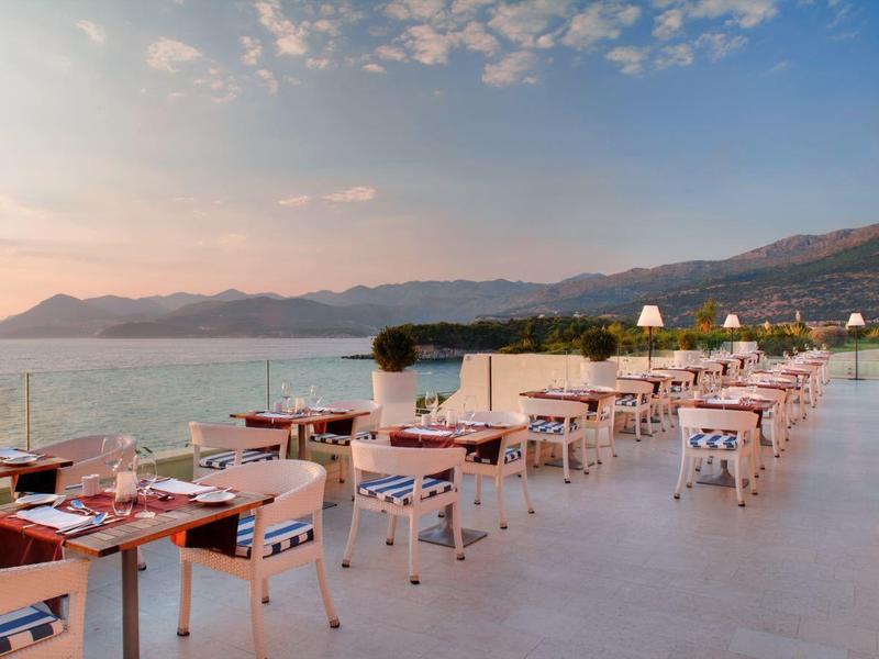 Velká terasa s stoly a židlemi s výhledem na moře a hory při západu slunce.