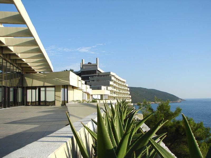 Modern hotel complex with terrace overlooking sea and green hills.