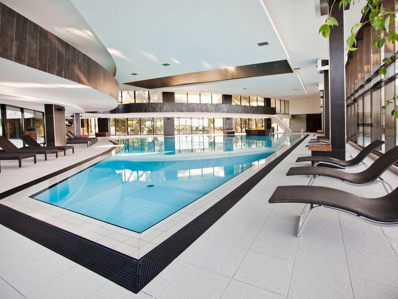 Modern indoor hotel pool with lounge chairs and large windows.