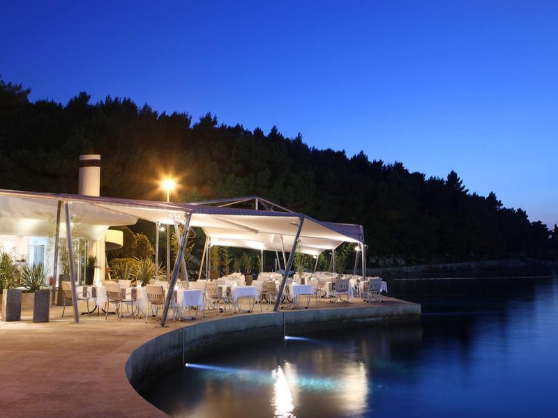 Evening view of a lit terrace by the lake with tables and chairs next to a forest.
