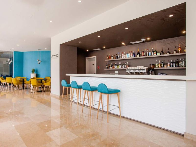 Moderní hotelový bar s modrými barovými židlemi a žlutými židlemi v světlém prostoru.