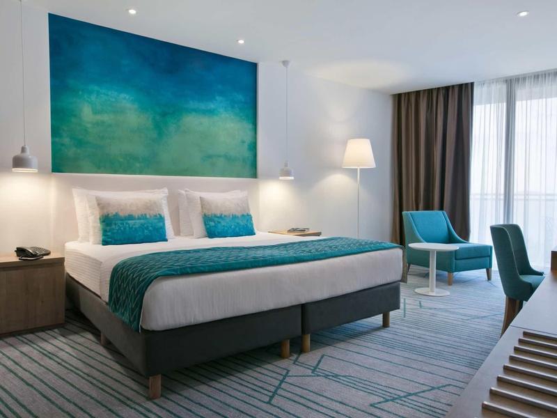 Moderní hotelový pokoj s velkou postelí, tyrkysovými prvky a velkým obrazem na stěně.