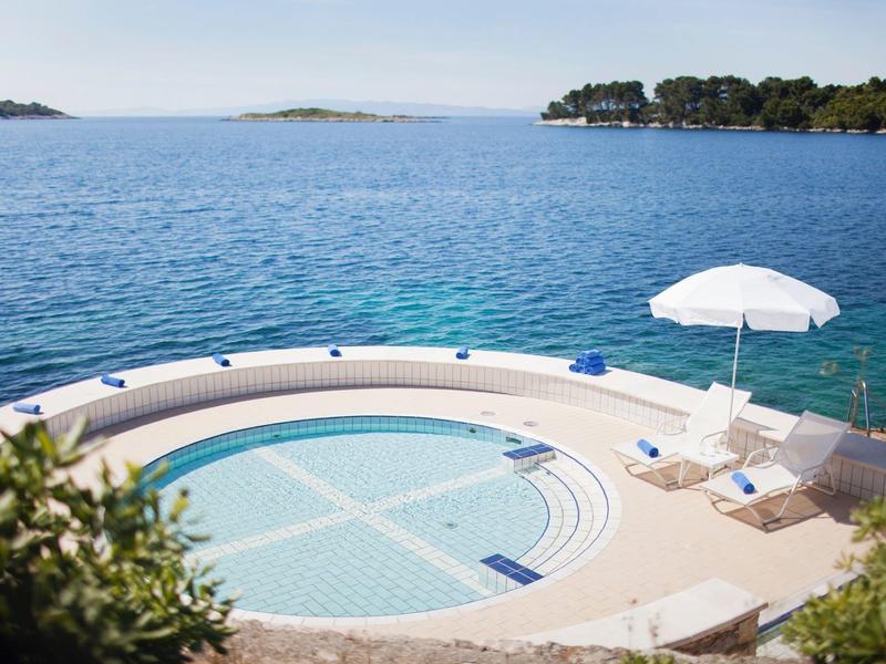 Okrągły basen infinity z widokiem na morze, leżaki i parasol przeciwsłoneczny.