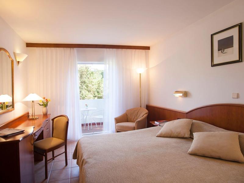 Jasny pokój hotelowy z łóżkiem podwójnym, biurkiem i balkonem z widokiem na zieleń.