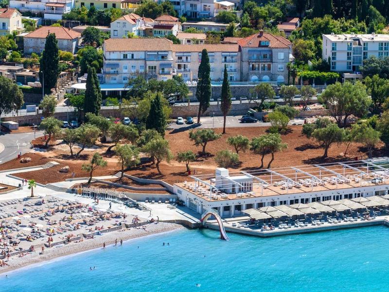 Strandresort met ligstoelen en helder blauw water voor een beboste heuvel met gebouwen.