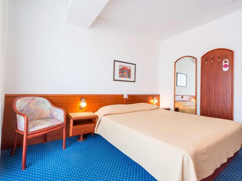 Licht ingericht hotelkamer met tweepersoonsbed, fauteuil, nachtkastje en blauw tapijt.