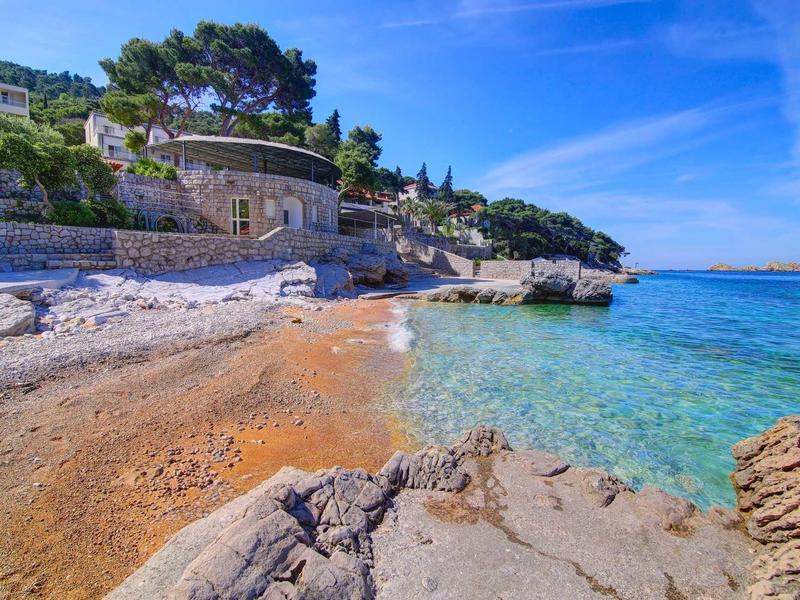 Plage de galets avec eau turquoise claire et maisons sur une falaise verte sous un ciel bleu.