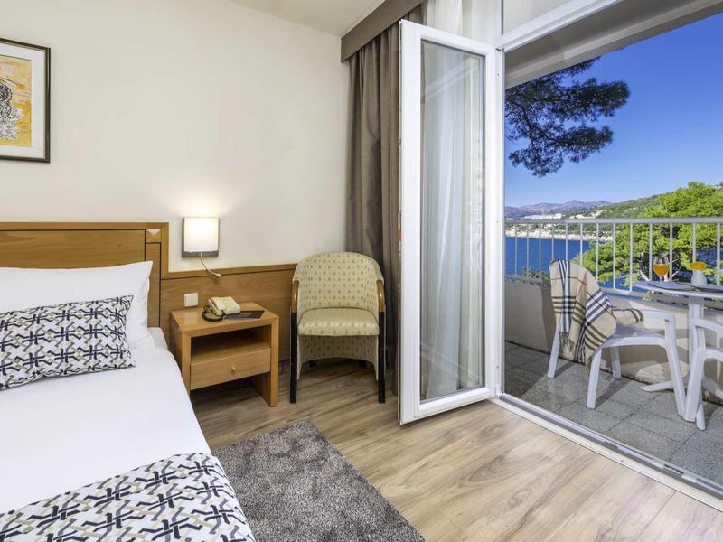 Chambre d'hôtel avec balcon, vue sur la mer, lit, fauteuil et table avec chaises.