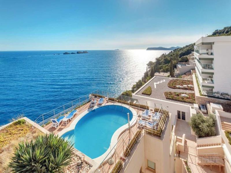 Meerblick mit klarem blauem Wasser, Pool, weißem Hotelgebäude und Grünflächen unter strahlendem Himmel.