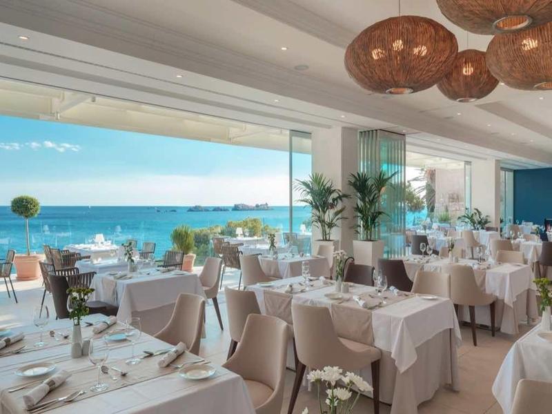 Elegantes Restaurant mit weißen Tischen, großen Fenstern zum Meer und modernen Kupferlampen.