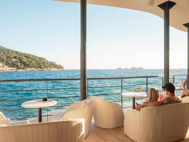 Balkon mit bequemen Sesseln und Tischen, Blick auf Meer und bewaldeten Hügel bei klarem Himmel.