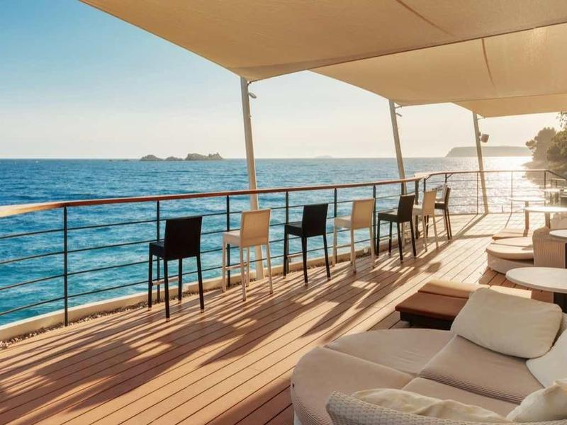 Terrasse mit weißen Sitzkissen, Stühlen und Tischen am blauen Meer unter Sonnenschutz.