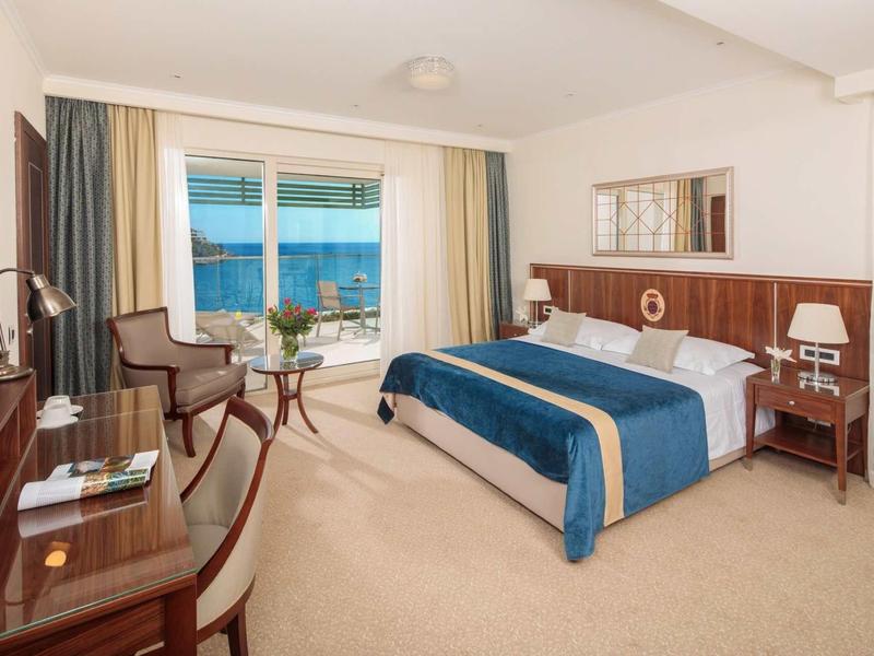 Hell eingerichtetes Hotelzimmer mit großem Bett, Schreibtisch, Stühlen und Blick aufs Meer durch Fenstertür.