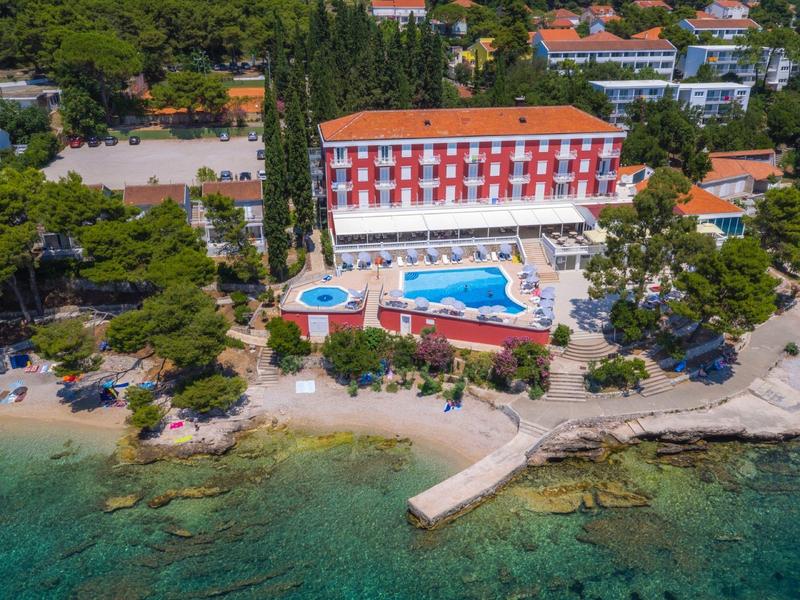 Beach hotel with pool surrounded by trees, located directly by turquoise sea with pier.