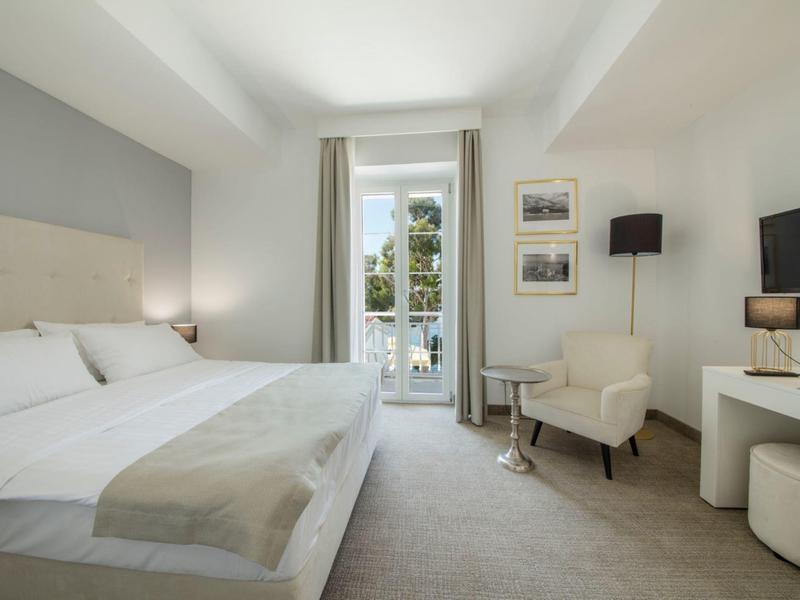 Modern hotel room with double bed, armchair, TV, and balcony overlooking trees.