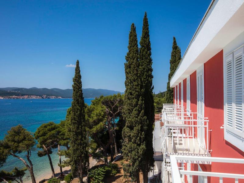 View from a hotel balcony of the sea and tall cypress trees under a clear sky.