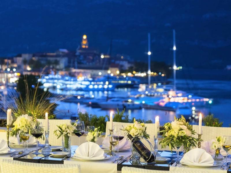 Elegant gedekte tafels met bloemen en kaarsen uitkijkend over verlichte haven 's nachts