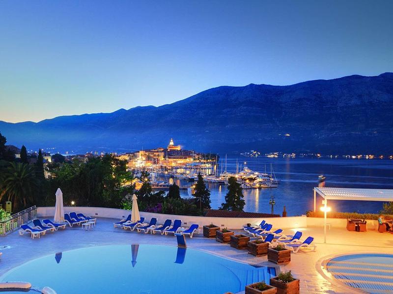 Nachtzicht op verlicht hotelzwembad met ligstoelen en haven op de achtergrond.