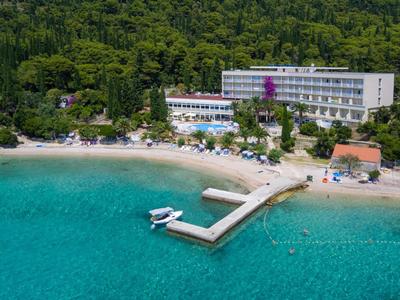 Klares türkisfarbenes Meer vor Strand mit Steg, Boot, Hotel und dichtem grünen Wald im Hintergrund.