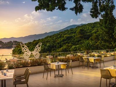 Terrasse eines Hotels mit Tischen und Stühlen vor einer Landschaft mit See und Bergen im Sonnenuntergang.