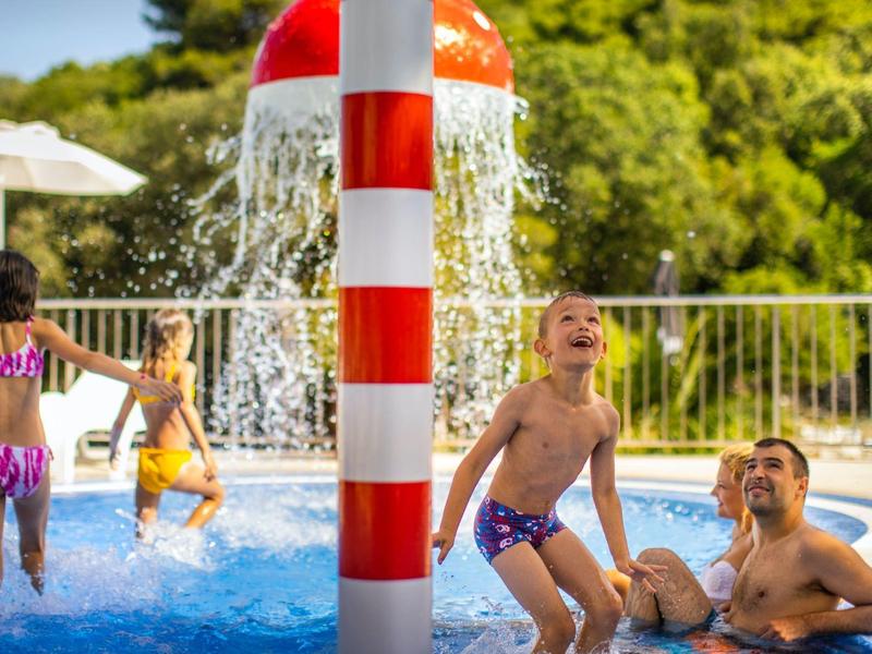 Kinder spielen und planschen unter einem Wasserspiel im Poolbereich im Freien.