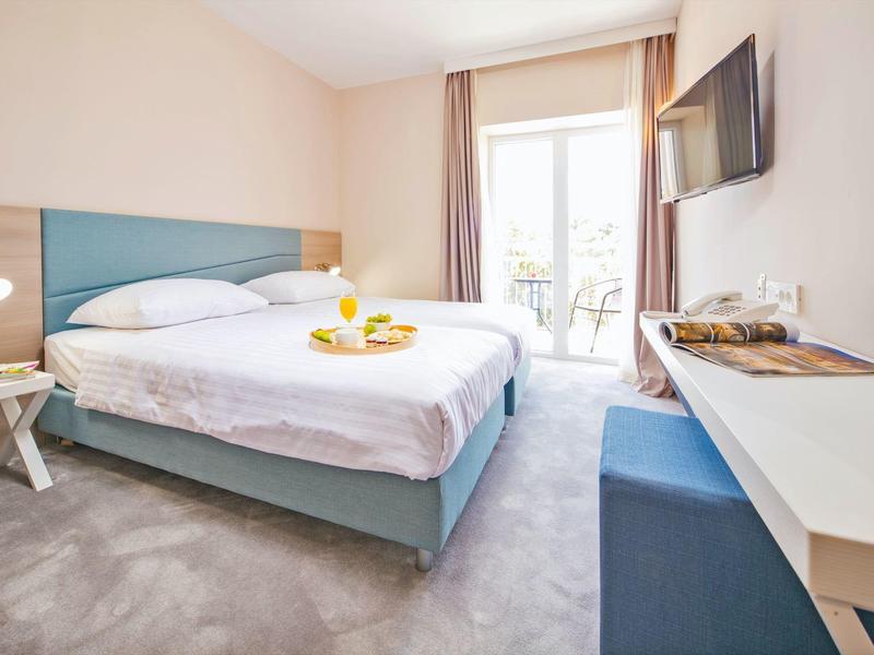 Helles Hotelzimmer mit Doppelbett, Fernseher, Schreibtisch und Balkon.