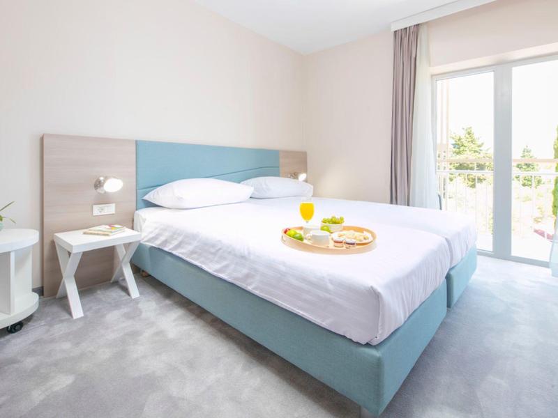 Helles Hotelzimmer mit blauem Bett, weißer Bettwäsche, Nachttischen und Balkon mit Tageslicht.