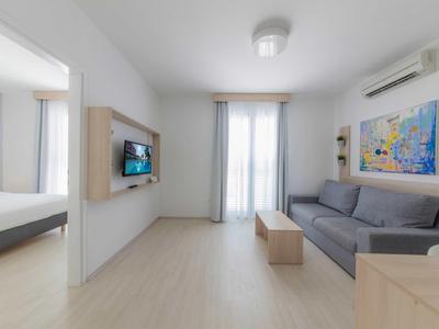 Lichte woonkamer met grijze bank, muurschildering en uitzicht op slaapkamer met tweepersoonsbed.