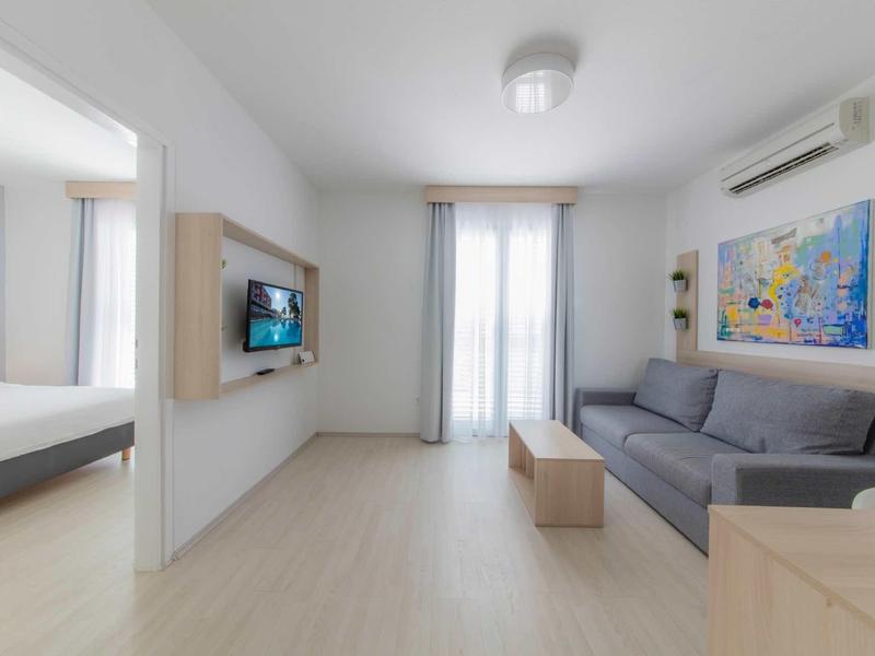 Lichte woonkamer met grijze bank, muurschildering en uitzicht op slaapkamer met tweepersoonsbed.